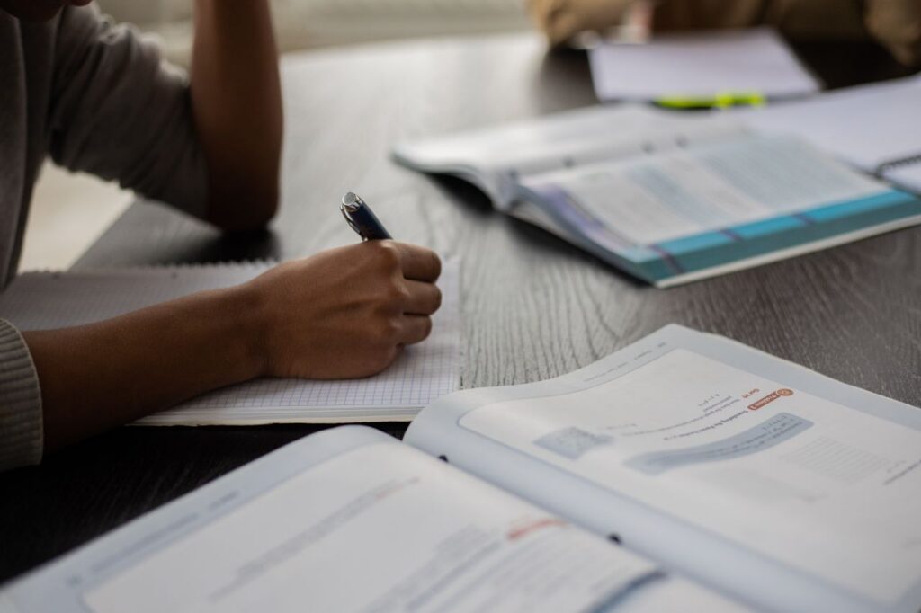 Side view of unrecognizable African American student sitting at table with textbooks while taking note in copybook in classroom with anonymous classmates on blurred background